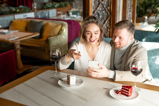 Giovane donna felice che indica allo schermo dello smartphone mentre discute qualcosa con il suo fidanzato da tavola servita nel ristorante