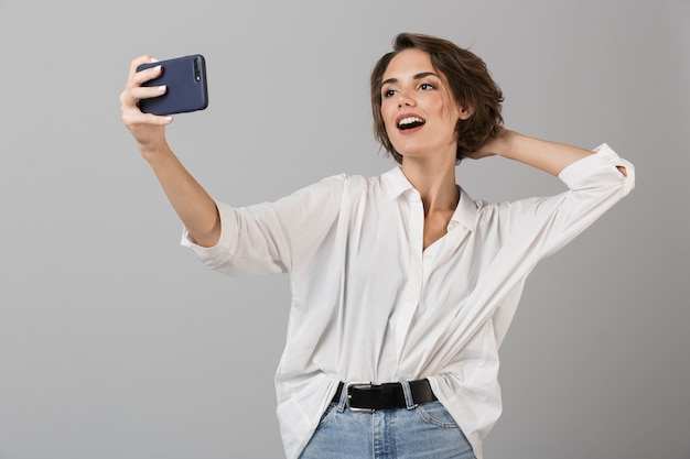 Happy young woman posing isolated over grey wall make selfie by mobile phone