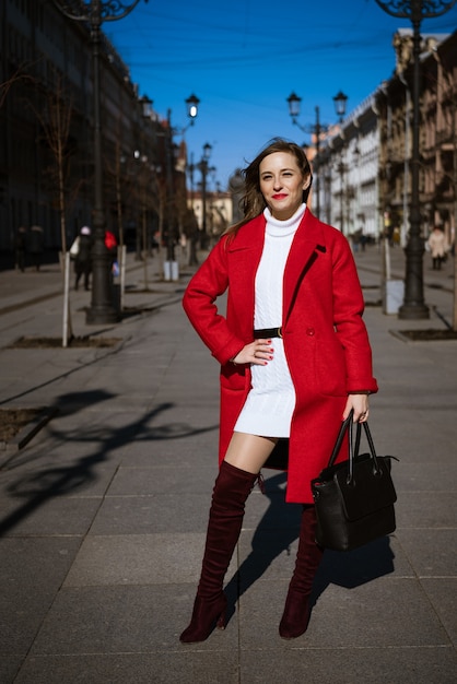 Happy young woman posing on the street in red coat with bag in hand