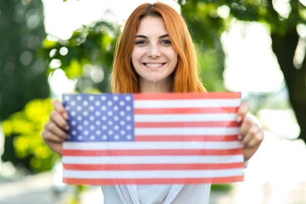 Счастливая молодая женщина позирует с национальным флагом сша на открытом воздухе в летнем парке. красивая девушка празднует день независимости соединенных штатов.