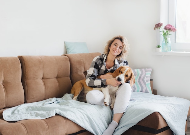 Happy young woman putting down a Beagle dog while sitting on a sofa in a checked shirt.