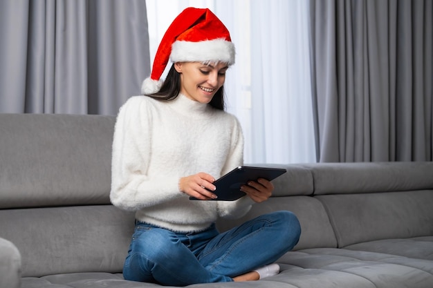 Happy young woman in a red Santa Claus hat sitting on the sofa and uses a tablet