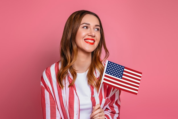 Happy young woman in a red shirt with red lipstick holds a small american flag and smiles isolated over pink space, USA flag, 4th of july independence day