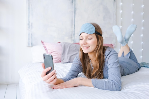 Happy young woman relaxing using and talking with smartphone on\
bed at home