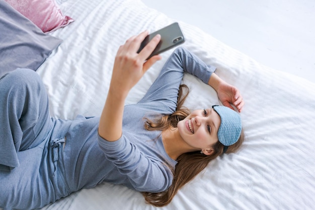 Happy young woman relaxing using and talking with smartphone on bed at home