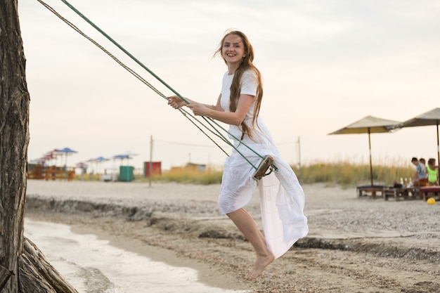 Счастливая молодая женщина едет на качелях веревки на пляже. Отпуск.