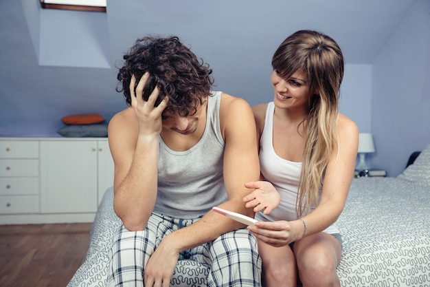 写真 ベッドの上に座っている心配している男性に妊娠検査を示す幸せな若い女性