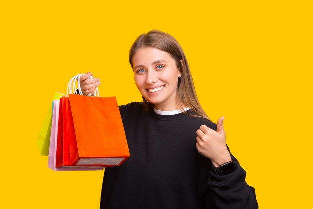 Happy young woman showing thumb up, holding shopping bags, looking excited at camera, yellow background