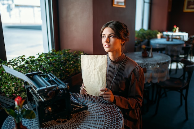 幸せな若い女性はカフェのテーブルのそばに座って、思慮深く窓の外に見えます。