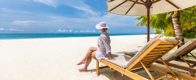 Happy young woman sitting on beach chair on tropical beach in white dress. Enjoying life, carefree