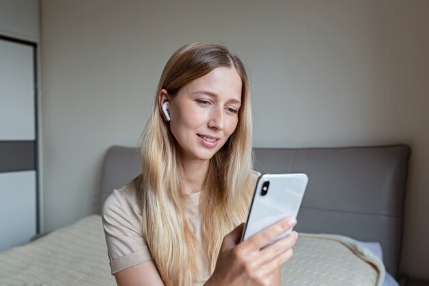 Happy young woman sitting on bed at home. Holding phone in hands and smile. Listening to music