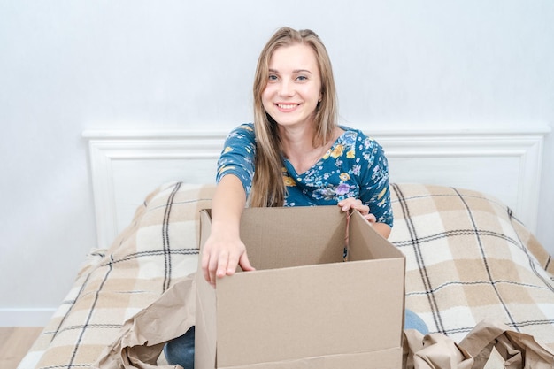 Happy young woman sitting on bed with open carton box at home Delivering parcel unpacking