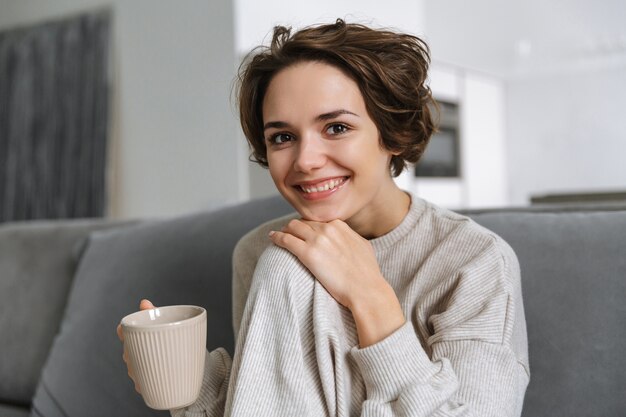 マグカップを持って、自宅のソファに座って幸せな若い女性