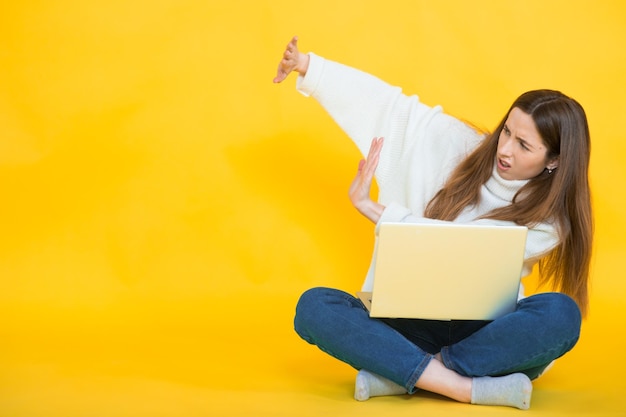 Happy young woman sitting on the floor with crossed legs and\
using laptop on yellow