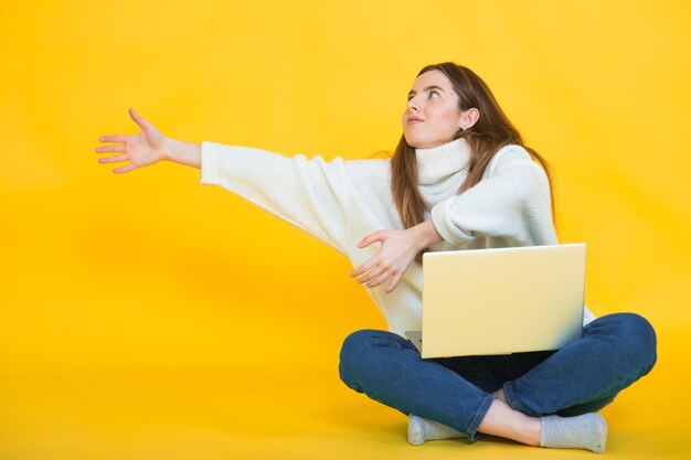 Happy young woman sitting on the floor with crossed legs and\
using laptop on yellow