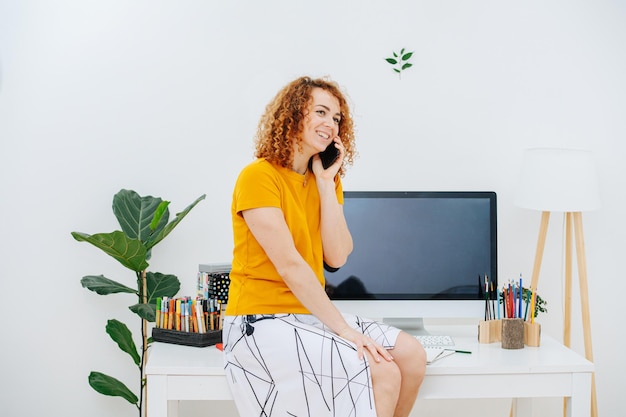 Happy young woman sitting on her workplace desk speaking on her phone