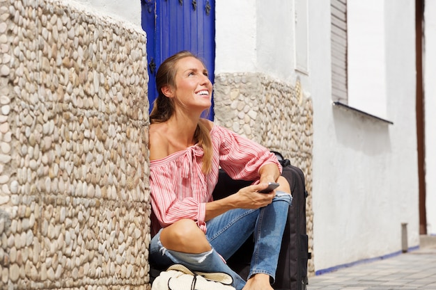 Happy young woman sitting on sidewalk with suitcase and cellphone