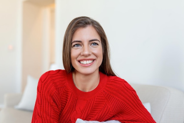 Photo happy young woman sitting on sofa at home and looking at camera portrait of comfortable woman in winter clothes relaxing on armchair portrait of beautiful woman smiling and relaxing during autumn
