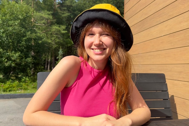 夏の日にパナマ帽子をかぶり、カメラを見て笑顔で屋外のテーブルのテラスに座っている幸せな若い女性