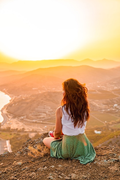 A happy young woman in a skirt is sitting on the top of a mountain cliff under the sunset sky