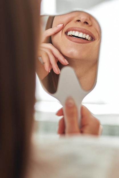 행복한 젊은 여성이 치과의사 사무실에서 거울에 비친 완벽한 건강한 치아를 보며 웃고 있습니다.