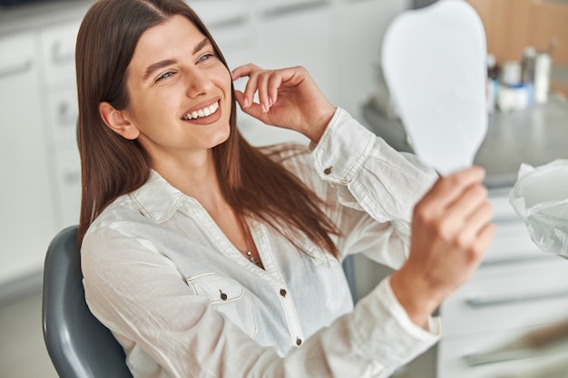 Felice giovane donna sorridente controllando i suoi denti sani perfetti allo specchio, seduta su una poltrona del dentista presso l'ufficio del dentista.