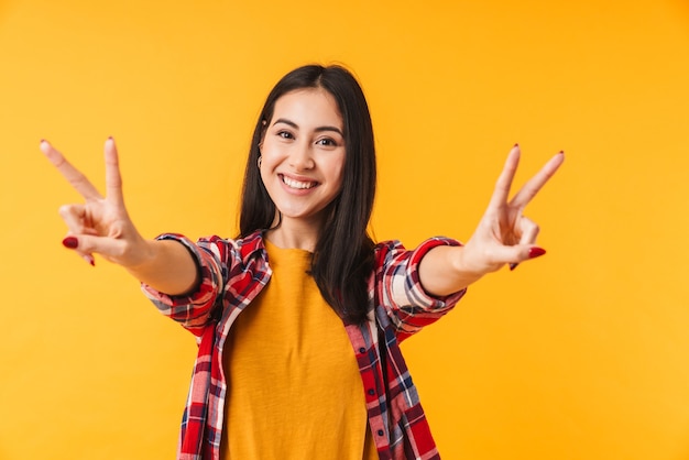 happy young woman smiling and gesturing peace sign isolated over yellow wall