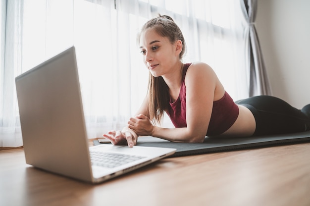 Felice giovane donna in abiti sportivi sdraiato sul pavimento utilizzando il computer portatile per imparare la lezione di allenamento online a casa