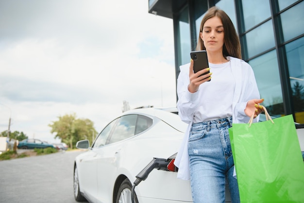 Felice giovane donna in piedi sul parcheggio della città vicino a un'auto elettrica che carica la batteria dell'automobile dalla piccola stazione della città che tiene le borse della spesa