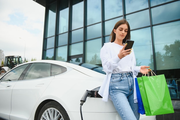 Happy young woman standing on city parking near electric car
charging automobile battery from small city station holding
shopping bags