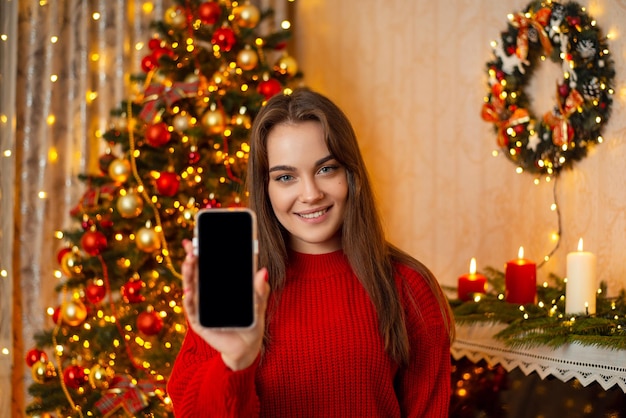 クリスマス ツリーの前に立っている幸せな若い女性はカメラにスマート フォンを示しています