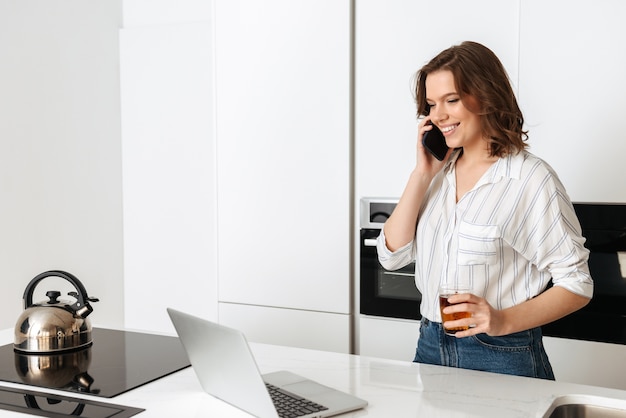 Happy young woman standing at the kitchen at home, using mobile phone