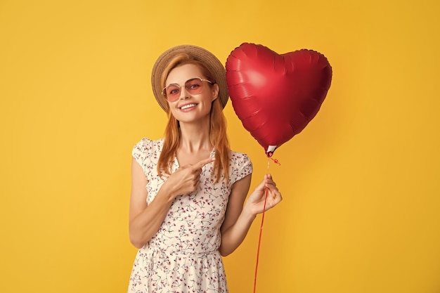 Happy young woman in straw hat and sunglasses point finger love heart balloon on yellow background