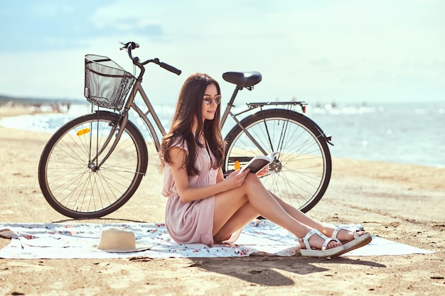 サングラスをかけた幸せな若い女性は、ビーチに座って本を持ってカメラを見ています。