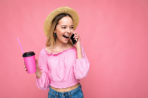 ピンクの背景にコーヒーを飲みながら携帯電話で話している幸せな若い女性。