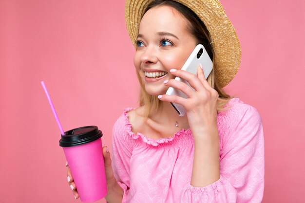 Happy young woman talking on mobile phone while having coffee on pink background.