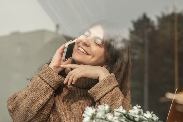 通りからの眺め、カフェに座って電話で話している幸せな若い女性。