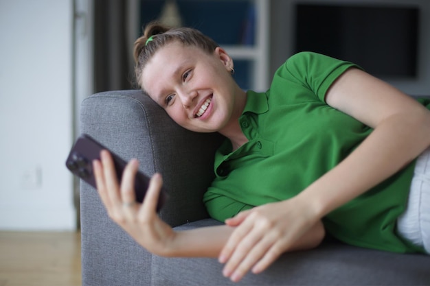 Happy young woman teenager girl lay on couch at home in living room and use cell mobile phone