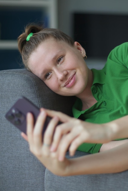 Happy young woman teenager girl lay on couch at home in living room and use cell mobile phone