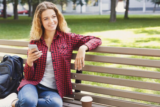 スマートフォンで幸せな若い女性のテキストメッセージ、ラップトップで作業しているフリーランサー、公園で屋外に座っています。テクノロジー、コミュニケーション、教育、リモートワークのコンセプト、コピースペース