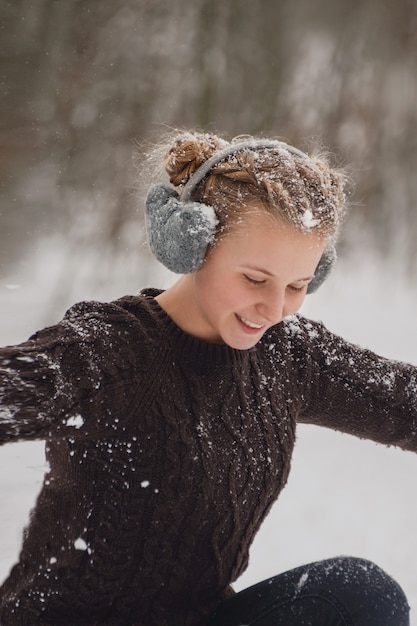 雪を投げる幸せな若い女性ふわふわのヘッドフォンとニットのセーターで凍るような冬を楽しんでいる美しい冬の女性公園屋外空飛ぶ雪片良い気分冬のクリスマス休暇のコンセプト