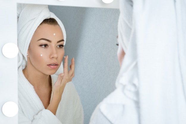 Felice giovane donna in un asciugamano davanti allo specchio applica la crema per il viso, un concetto di cura della pelle a casa