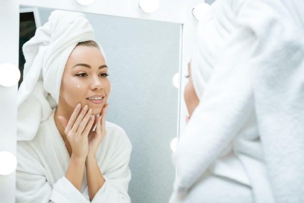 A happy young woman in a towel in front of a mirror applies cream to her face, a concept of skin care at home