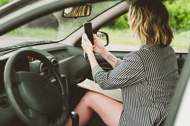 幸せな若い女性が道路を車で旅行します彼女は地図と電話を見て