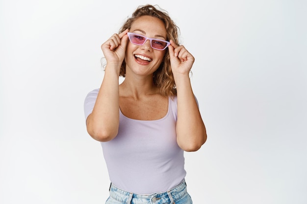 Happy young woman tries new sunglasses, smiles and looks pleased at camera, stands in stylish summer outfit against white background.