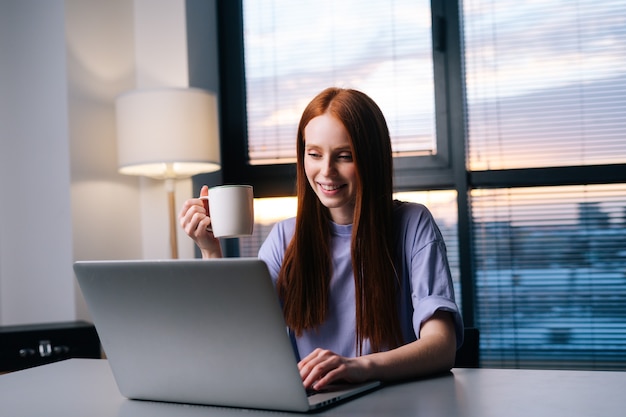 Happy young woman using laptop computer and drinking coffee at home workplace.