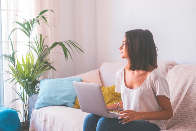 Happy young woman using laptop while sitting on sofa and looking out through window from the living room of her house. Thoughtful lady spending leisure time using laptop. Freelancer working from home