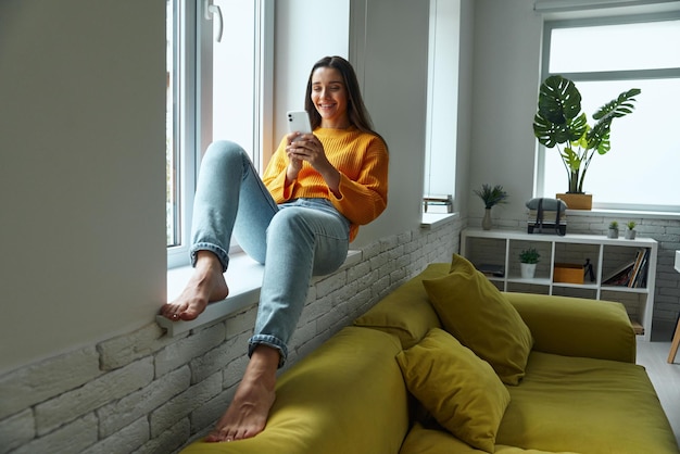 Photo happy young woman using smart phone while sitting on the window sill at home