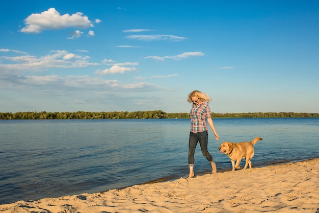 Happy young woman walking along a beach with her golden retriever at the sunset