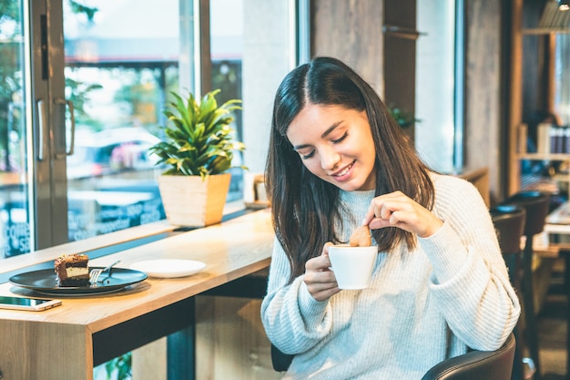 Happy young woman in a warm sweater with cup of coffee and a biscuit sitting by a window